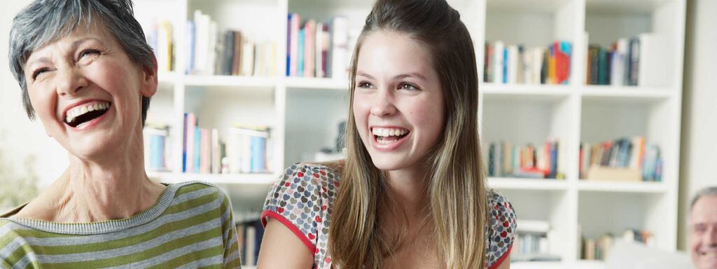 A smiling older woman and a teenage girl share a joyful moment together in a cozy, well-lit room filled with books.