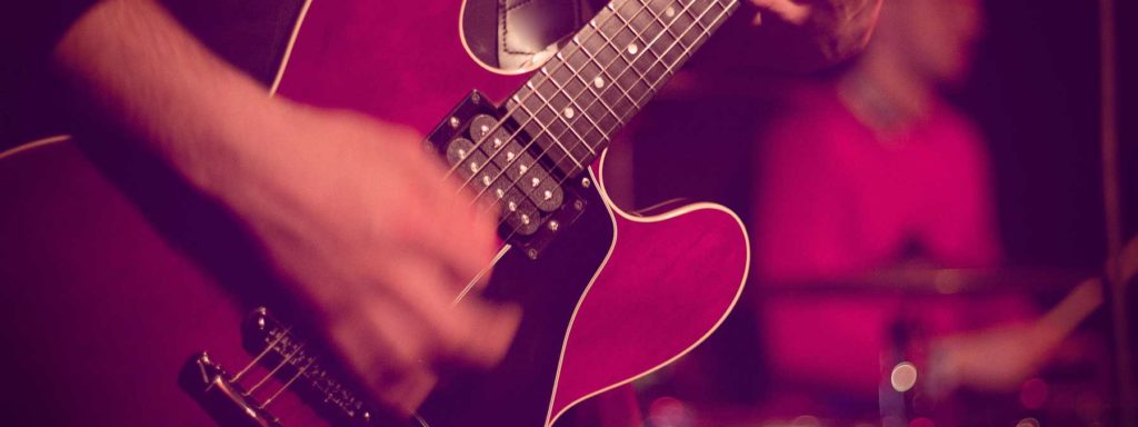 A close-up of a hand playing a guitar, with a blurred drummer in the background. The guitar is dark-colored.