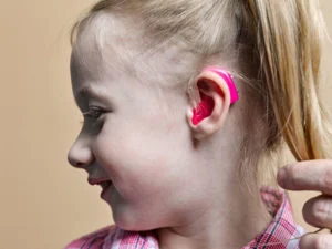 Child with a pink hearing aid in her ear, smiling while a caregiver adjusts the device.
