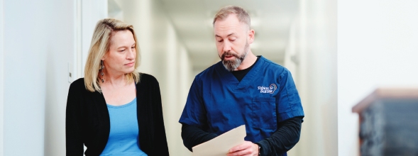 An audiologist consults with a female patient, discussing information on a sheet of paper in a hallway setting.