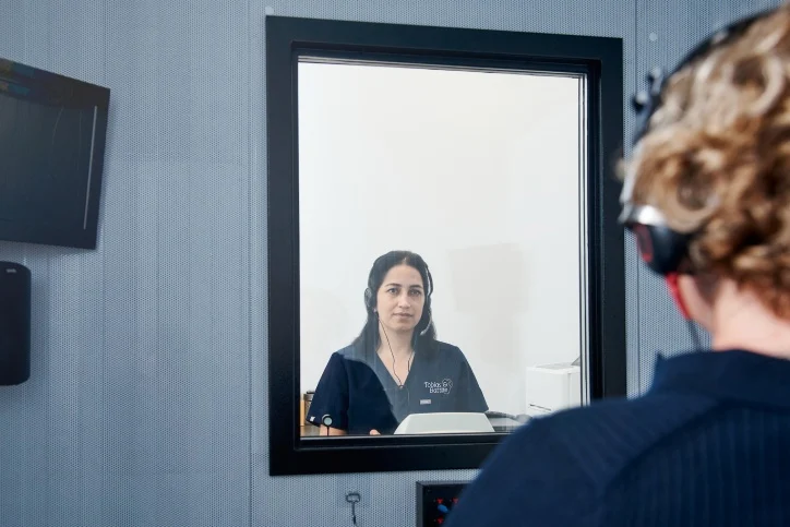 An audiologist tests a patient's hearing in a soundproof room, with the patient wearing headphones and facing a glass window.
