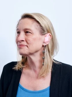 A woman fitting custom ear protection in her ear, wearing long earrings and a blue top.
